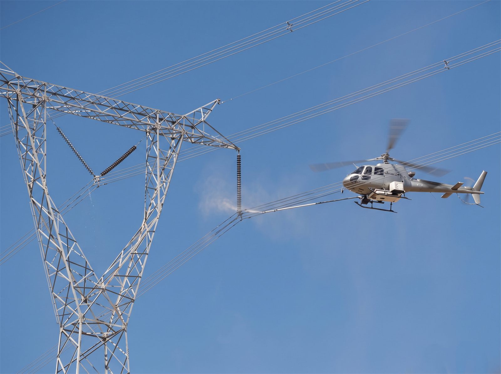 Helicopter hovering beside a transmission tower, washing an insulator string with a long spray lance